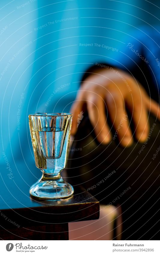 On a table is an old shot glass. The hand and arm of a person can be seen against a blue background. Schnaps glass Alcoholic drinks vodka alcoholism Alcoholics