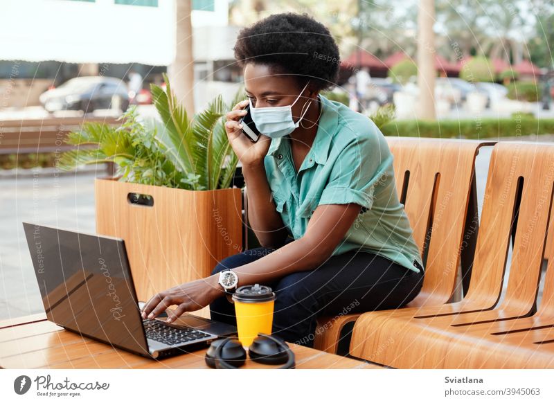 A young African girl in a medical face mask is talking on the phone and working with a laptop at a table in a cafe. Social distancing and work, work online, business online
