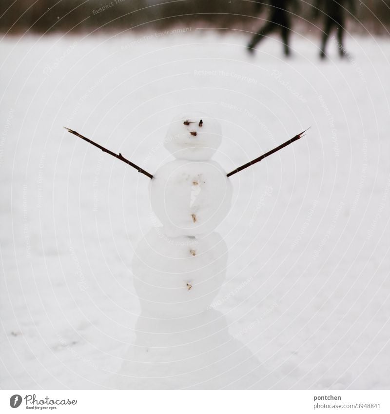 A snowman with outstretched arms stands in the snow. Blurred walkers in the background. Winter Snowman Children's game Joy fun pleasure Cold strollers Seasons