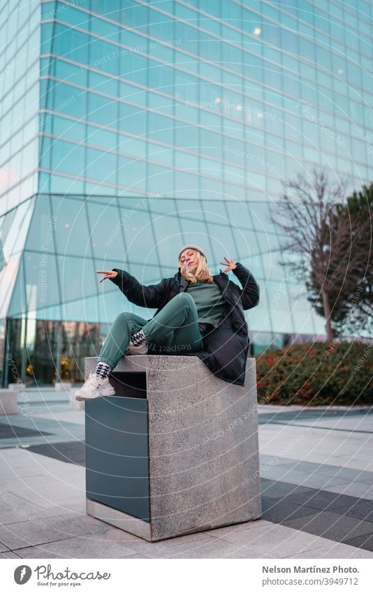 Blonde woman in a green outfit, a gray wool hat and a black jacket with a building in the background fashion portrait cold wear cute outdoor pretty apparel