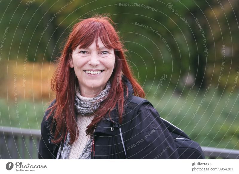 Happy smiling woman with red long hair outside in nature Looking into the camera Front view Upper body portrait Central perspective Shallow depth of field