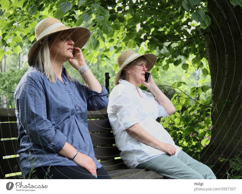 "It's about time we met again! I'm glad!" - Two women with hats sit on a bench and talk to each other on the phone. Human being Woman Senior citizen Bench Tree