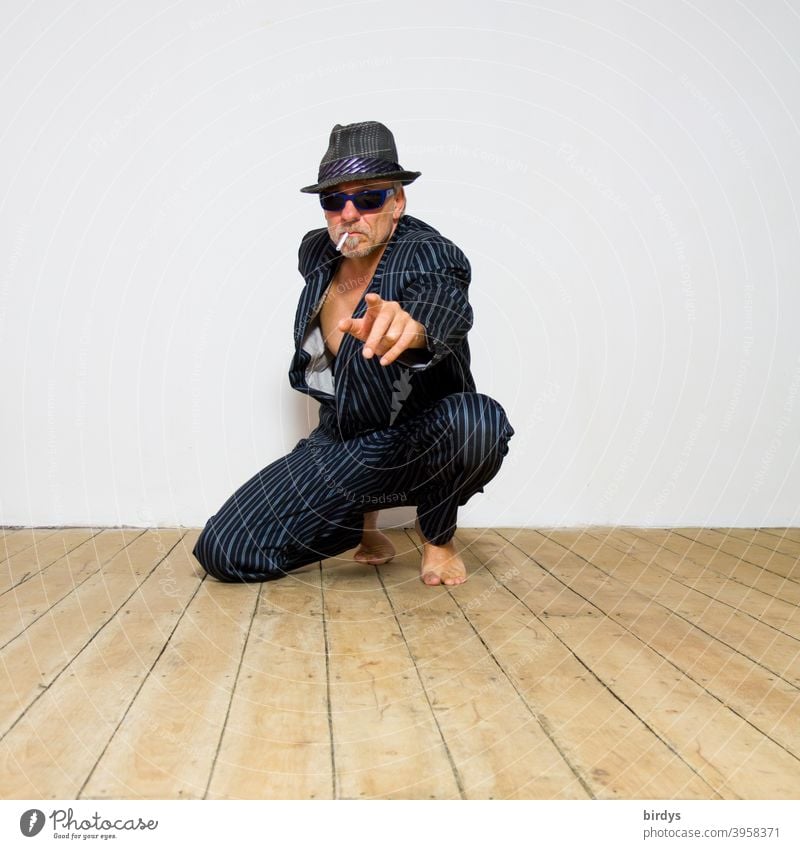 kneeling man with hat, sunglasses and cigarette points with outstretched arm and index finger into the camera. Barefoot in suit Man Cool Hat Sunglasses