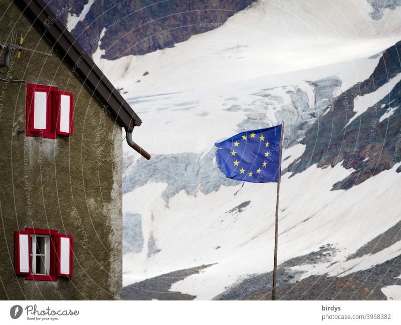Flag of the European Union in front of a house in the mountains , in the background a snow covered mountainside . EU - flag European flag Alps Snow