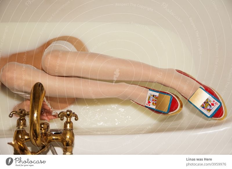 Long legs of a woman is the main subject of this image. She is relaxing in a milk bath with her red avant-garde shoes on. Golden cards and golden crane accompany her with these high fashion shoes.