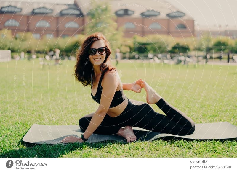 Happy fit young woman does stretching workout on fitness mat practices yoga outside dressed in activewear has strong body breathes fresh air outdoor leads active lifestyle. Sport fitness concept