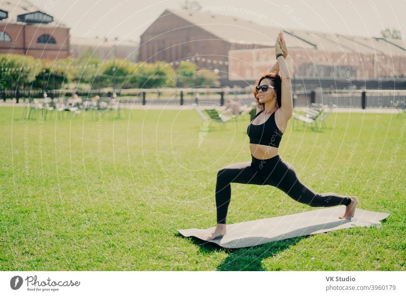 Concentrated positive woman raises arms does pilates exercises dressed in active wear sunglasses has workout during summer practices yoga poses on green grass. People and healthy lifestyle concept