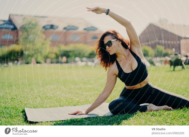 Image of motivated fit woman raises arm and does stretching exercises outdoors poses on fitness mat wears sunglasses and activewear trains actively. Fitness trainer goes in for sport outdoors