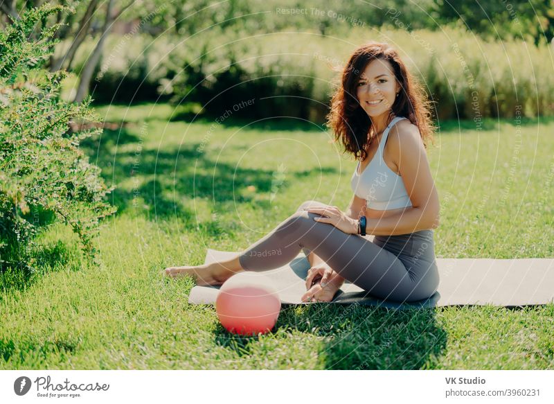 Positive brunette woman in cropped top and leggings poses bare feet on karemat uses ball for gymnastic exercises smiles pleasantly enjoys beautiful nature view. Sport outdoor. Yoga practice.