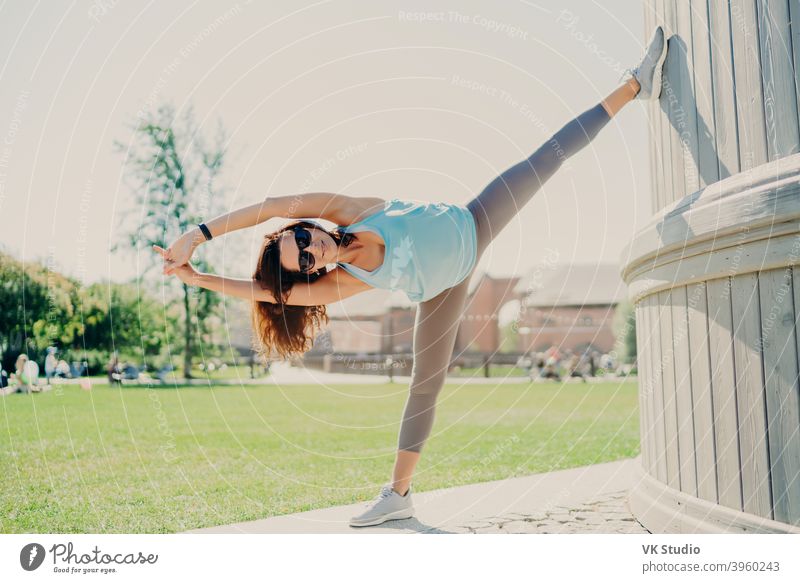 Fit brunette woman dressed in sportswear stands bare feet on karemat has  fintess training on green lawn outdoor during summer time wears sunglasses  looks somewhere being in good physical shape - a