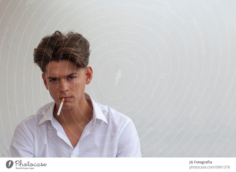 Young man with cigarette in his mouth looking at camera with gray wall background. young guy smoking portrait harmful cancer model freckled white caucasian