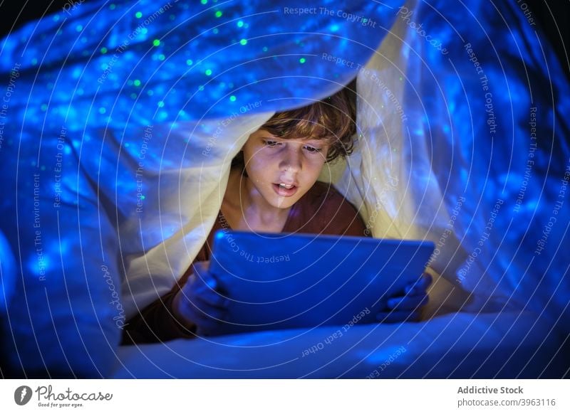 Content boy with tablet in dark room browsing kid cheerful bed bedroom blanket hide playful smile internet child happy device online gadget using sit chill