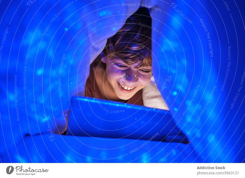 Content boy with tablet in dark room browsing kid cheerful bed bedroom blanket hide playful smile internet child happy device online gadget using sit chill