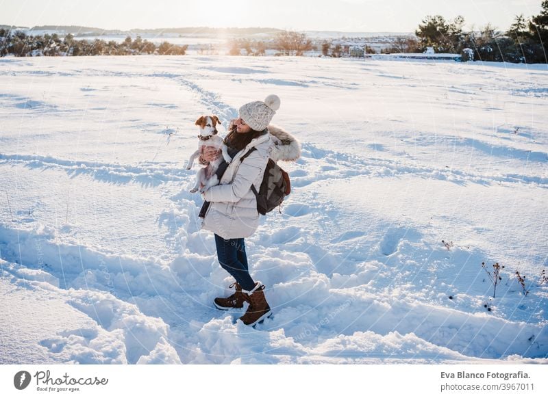 back view of woman in snowy mountain wearing modern coat at sunset. Holding cute jack russell dog in arms. winter season. nature at sunset travel owner pet love