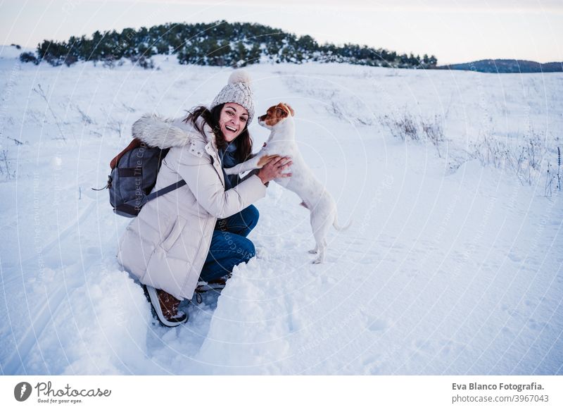 happy woman in snowy mountain wearing modern coat at sunset. Holding cute jack russell dog in arms. winter season. nature at sunset travel owner pet love