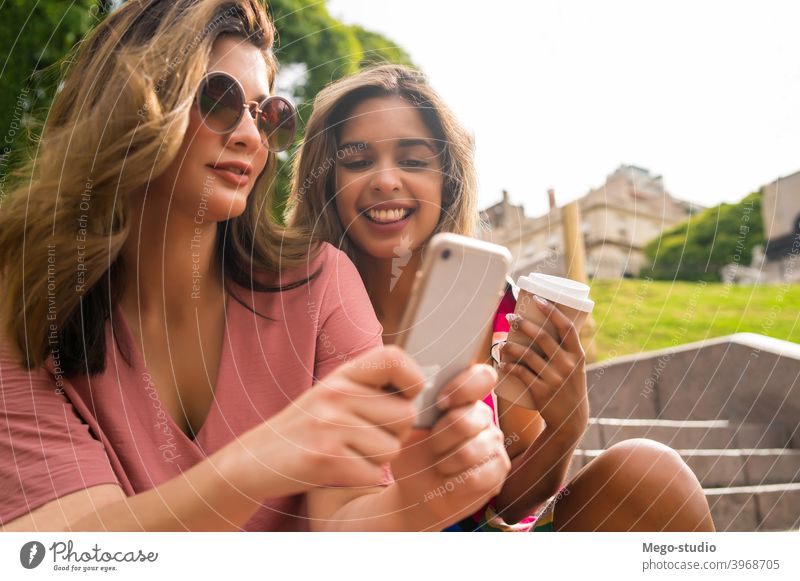 Two friends using mobile phone outdoors. two young smiling laughing friendship lifestyle leisure talking fun women cellular communicate app enjoying network
