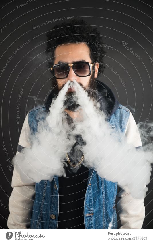 Young man vaping, studio shot. Bearded guy with sunglasses blowing a cloud of smoke on black background. Concept of smoking and steam without nicotine. beard