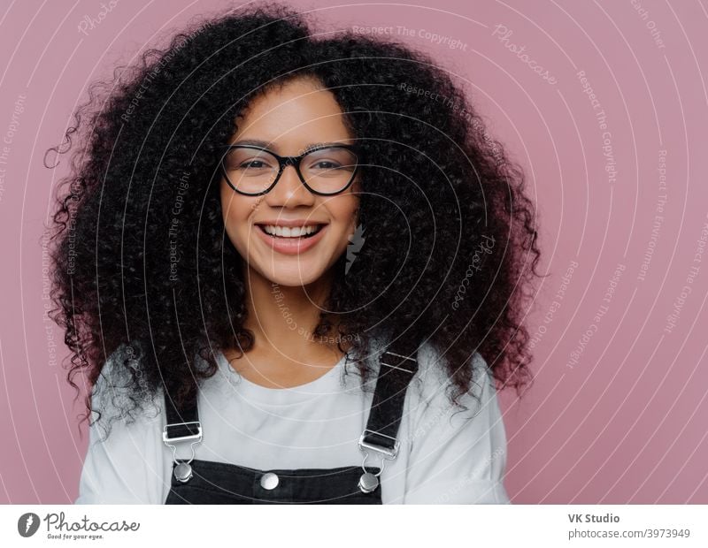 Close up portrait of dark skinned woman with crisp hair, smiles happily, wears optical glasses, casual clothes, poses over purple background expresses good emotions. Afro American student poses indoor