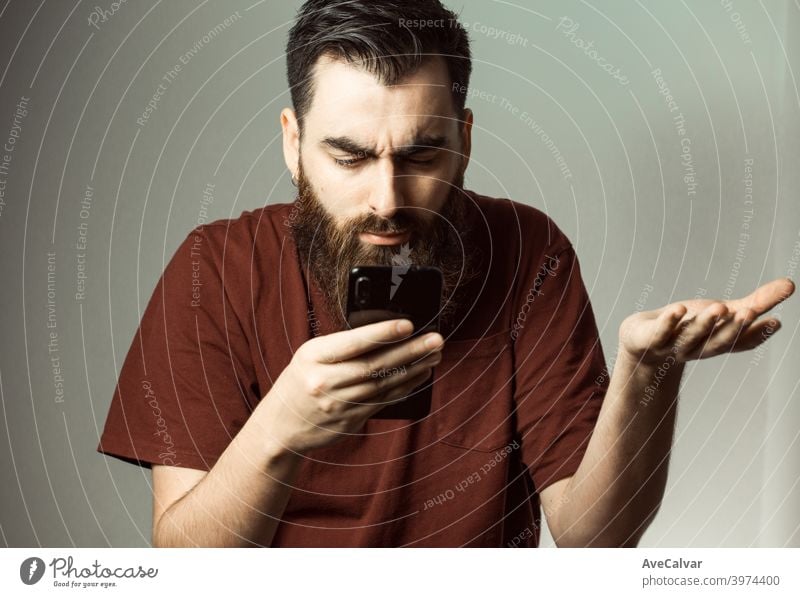 A young hipster style man with a beard and modern haircut angry while looking at his phone screen with copy space anxiety confused mistake anxious depressed