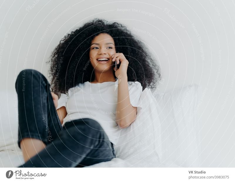 Pleasant looking curly woman has pleasant conversation, talks with friend via smartphone while relaxes in bed, has joyful expression, looks aside, wears casual clothing. People, bedding, technology