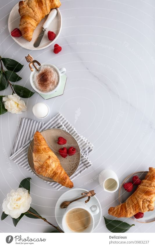 View from above of a breakfast table with croissants, coffee, raspberries, blood orange, boiled egg and flowers on a white wooden table, on white marble surface