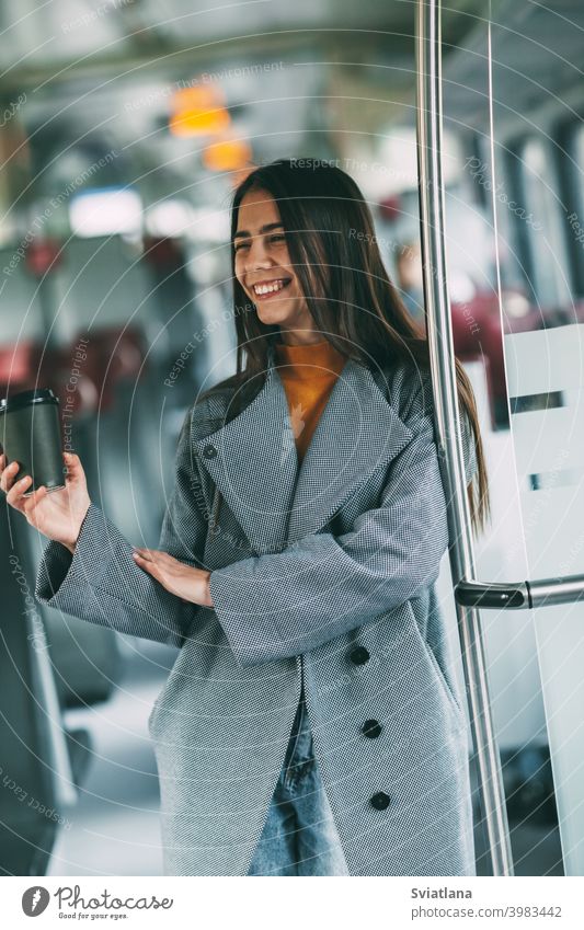 A happy brunette with coffee in her hands stands on the train, looks into the distance and smiles. transport young female beautiful railway passenger metro
