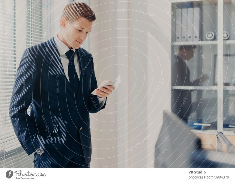 Concentrated male boss, keeps hand in pocket, focused into screen of smart phone, searches useful information for new startup, poses in office, dressed elegantly. Man chats on cellular at cabinet