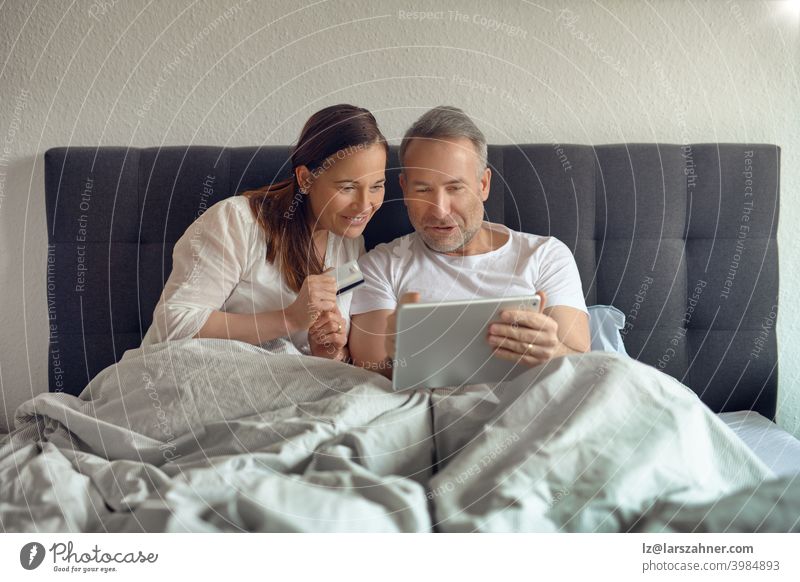 Happy middle-aged couple sitting in bed in the morning drinking coffee and doing online shopping with a digital tablet sofa furniture family room reclining