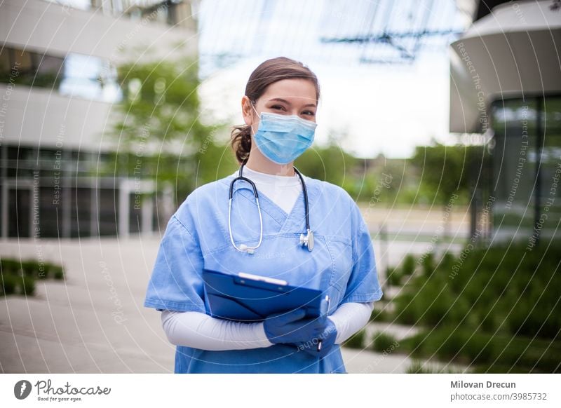 Doctor in front of hospital or nursing home, wearing blue scrubs, face mask, holding clipboard with patient form assisted living building care convalescent