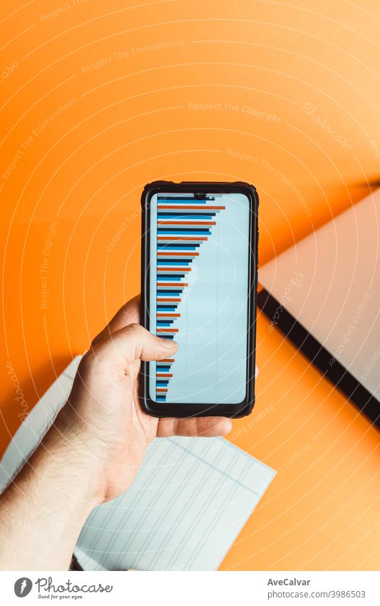 A hand holding a phone with statistics over a orange background with laptop and notebook with financial concept blank business office desk empty keyboard