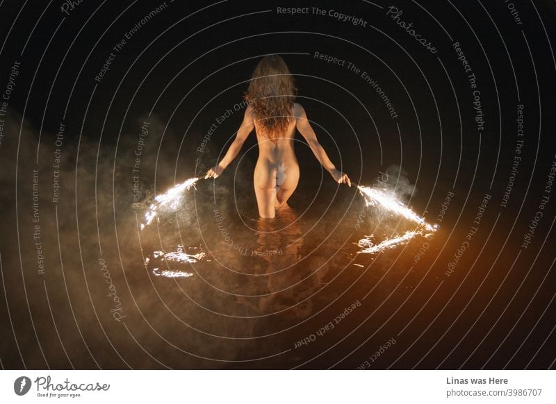 Wild and naked girl is swimming at night. Erotic image of a brunette model. Fireworks in her hands, sexy back show her perfect curves, smoke, and dark water makes this scene moody and sensual.