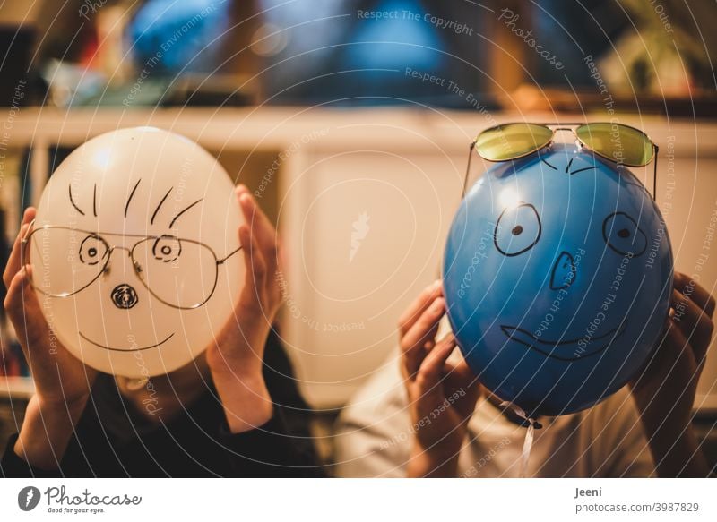 Balloons painted with funny faces in front of the heads of two children | Painted eyes, nose, mouth, hair and attached glasses | Hands hold the balloon tightly