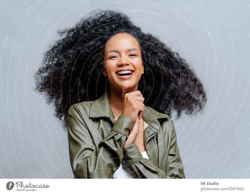 Overjoyed dark skinned curly woman laughs happily, laughs at funny joke, keeps hands pressed together, dressed in fashionable clothes, isolated over grey background. People and positiveness.
