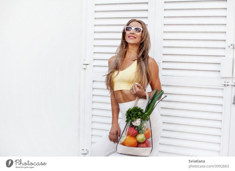 A young girl in sunglasses and sportswear stands in the background of the door with a bag full of vegetables and fruits. The concept of healthy eating and healthy lifestyle