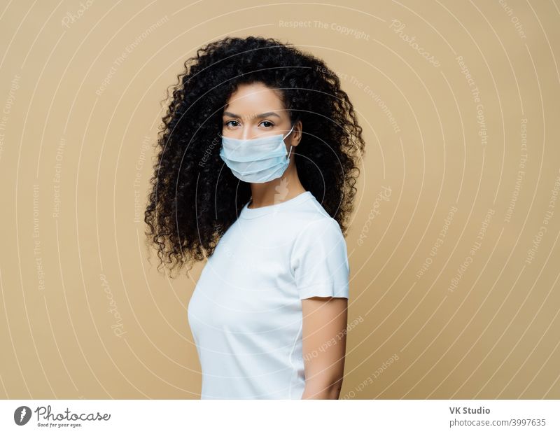 Sideways shot of Afro American woman wears protective face mask, protects against spread of coronavirus disease, stays at home, follows rules of self isolation. Covid-19, safety, quarantine concept
