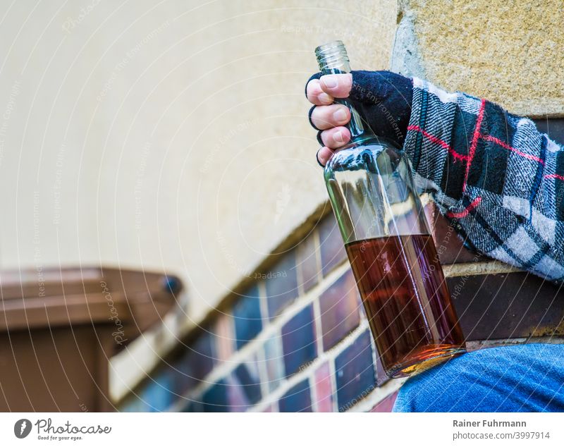 Close-up of an anonymous man sitting by a building and holding an open bottle of schnapps Alcoholic drinks alcoholism Addiction Building dustbin drunk Drinking