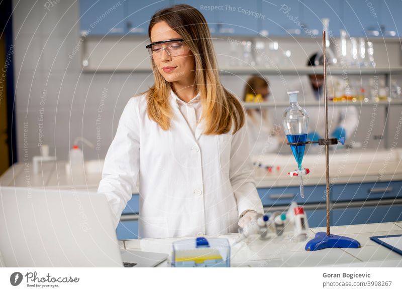 Female researcher in white lab coat and protective mask using laptop while working in the lab analyzing background bio chemical chemist clinic computer