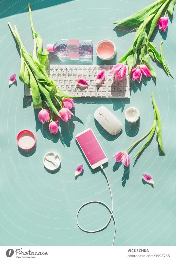 Female home office. Pastel blue desktop with PC keyboard , smartphone with blank screen and wire , cosmetics and pink tulips flowers in sunlight. Top view. Flat lay. Springtime. Blog layout