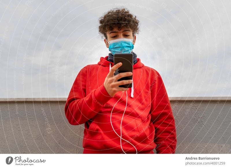 Young attractive man with curly hair dressed in a red sweatshirt using the mobile on the terrace of the house wearing a mask to protect himself from coronavirus