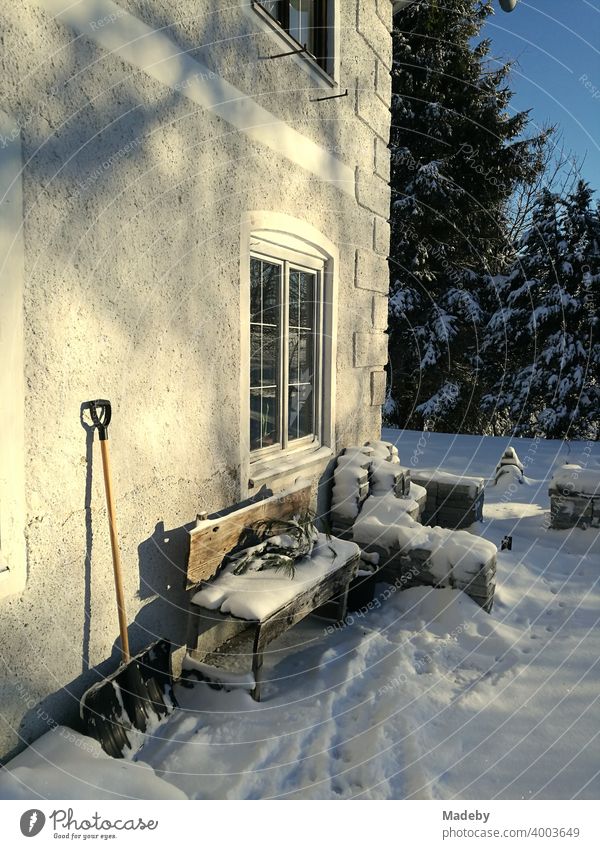 Snow-covered wooden bench and leaning snow shovel at an old farmhouse in winter at sunshine in romantic snowy landscape in Rudersau near Rottenbuch in the district Weilheim-Schongau in Upper Bavaria