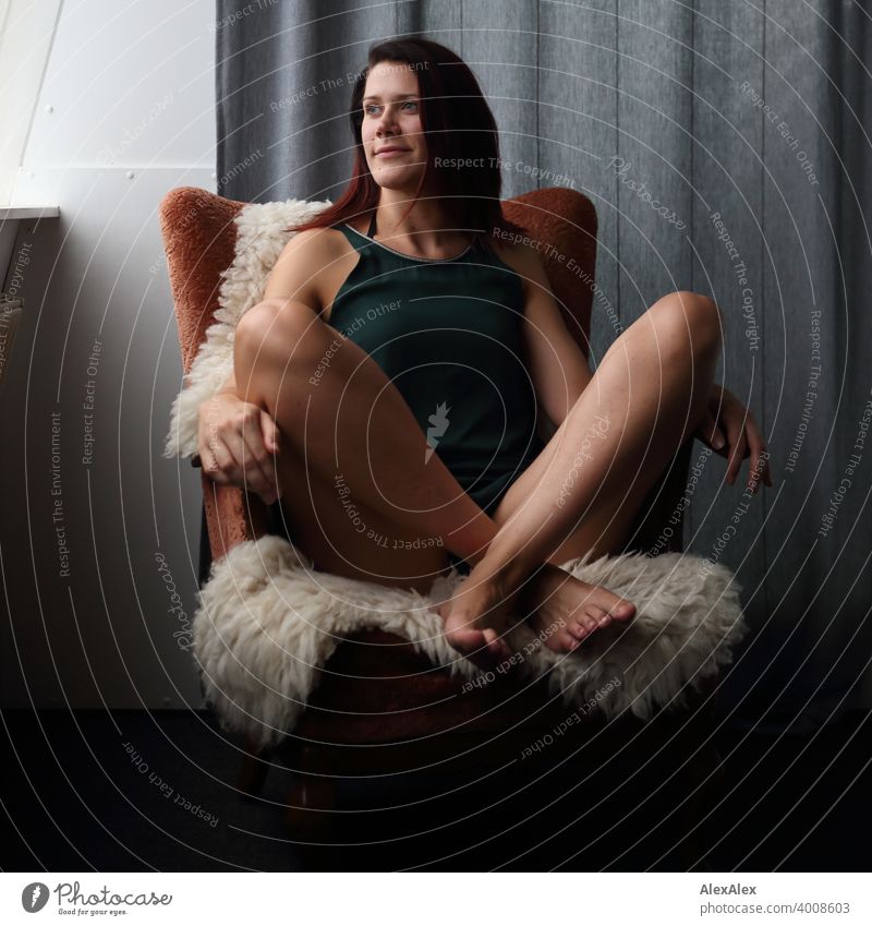 young woman sits cross-legged on an armchair with sheepskin and looks at the window Legs Barefoot Long-legged Laughter petrol Dress Drape Skin Colour photo