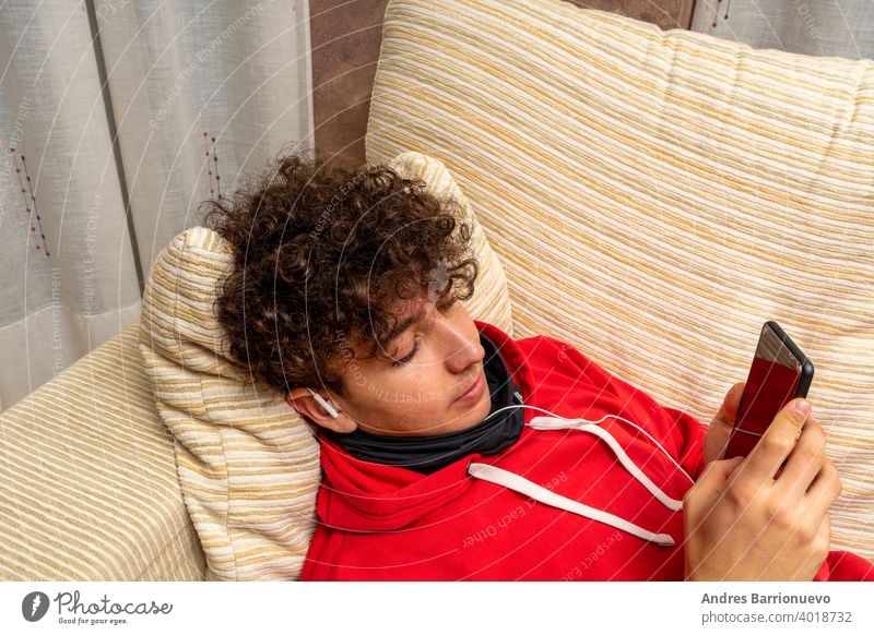 Young attractive man with curly hair dressed in a red sweatshirt using mobile in living room at home wearing a mask to protect himself from coronavirus