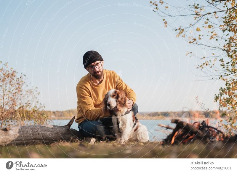 Man hugs his dog sitting by the lake. Active rest with pets, going on adventures, companionship: spaniel dog and his owner sit outdoors 25-35 years active
