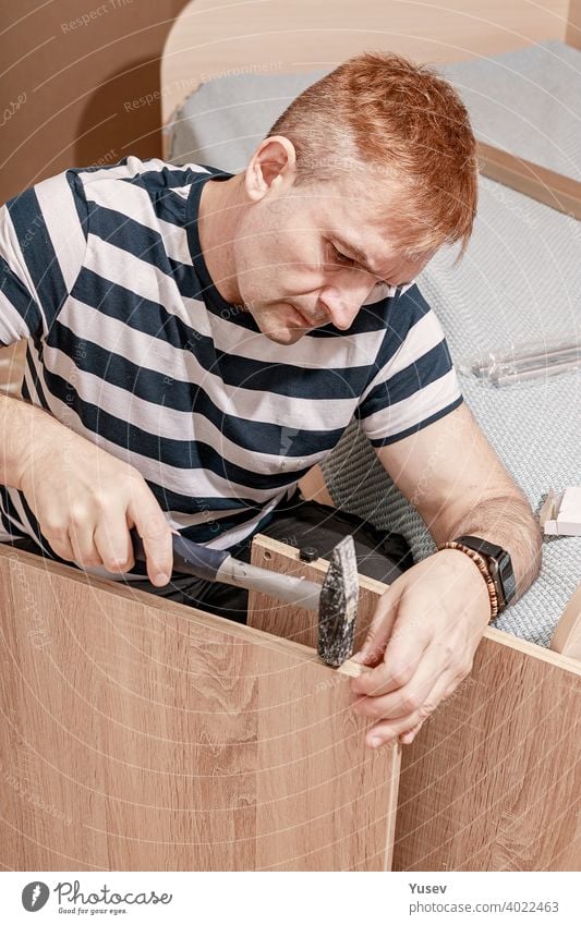 Good looking concentrated caucasian man in striped t-shirt assembles furniture with a hammer. Furniture assembly, repair, moving. Vertical shot good looking