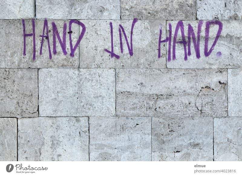 "HAND IN HAND" is written in purple block letters on the grey natural stone wall / together. Hold hands Wall (building) Facade in common Password Graffito