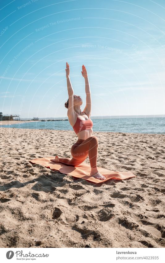 Young female in orange top and leggins practising Crescent Lunge Pose (Anjaneyasana) on a beach in the morning woman exercise pose yoga young practicing outdoor