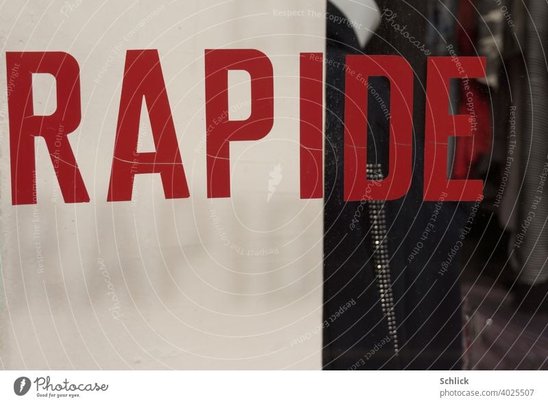 "RAPIDE" is written in French on a shop window in red capital letters, casting its drop shadow "RAPID" translated into German on a white background Text