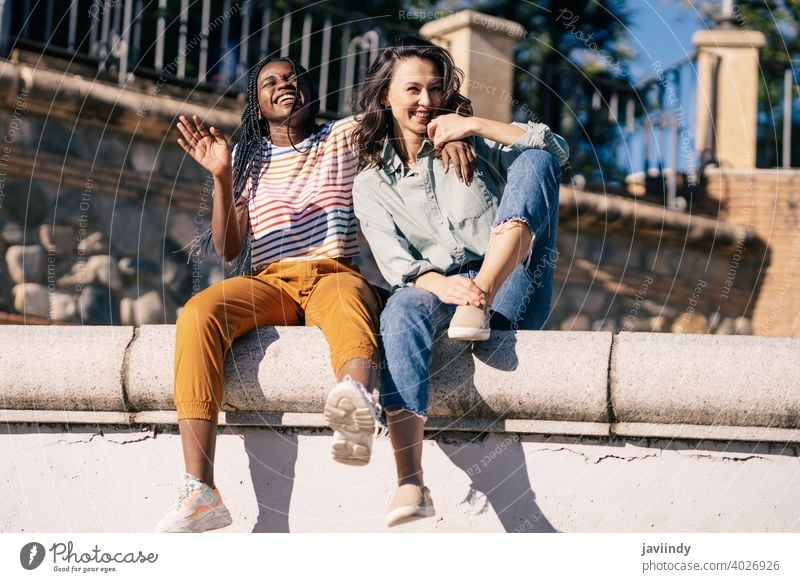 Two friends having fun together on the street sitting on a urban wall. women multiethnic black afro girl student two people lifestyle smile female pretty young