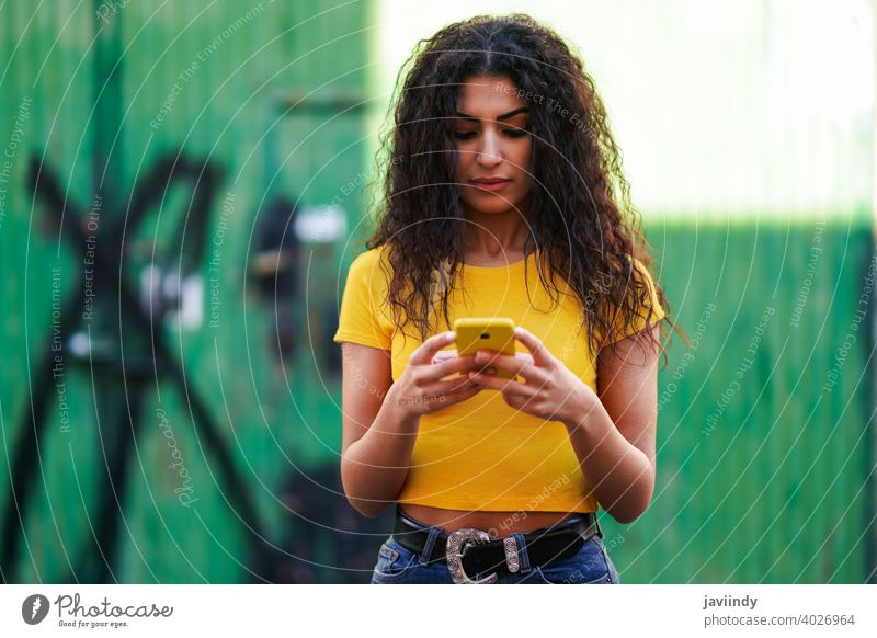 Young Arab woman walking in the street using her smartphone arab young texting message typing yellow copyspace girl recording hairstyle attractive beautiful