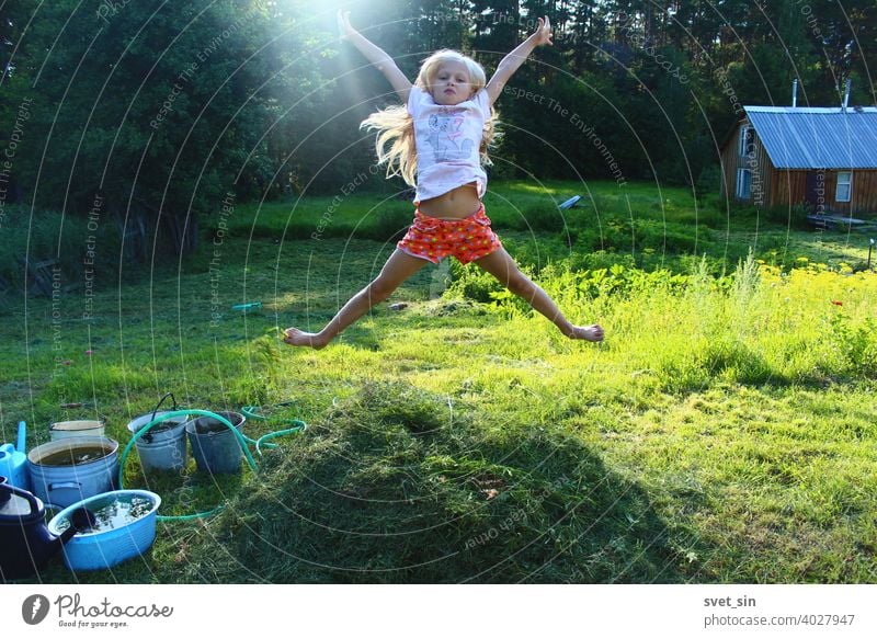 Barefoot childhood in the village. Portrait of a little blonde girl jumping in the air over a small stack of freshly cut grass on a green sunny lawn against the backdrop of a forest and a wooden bathhouse.